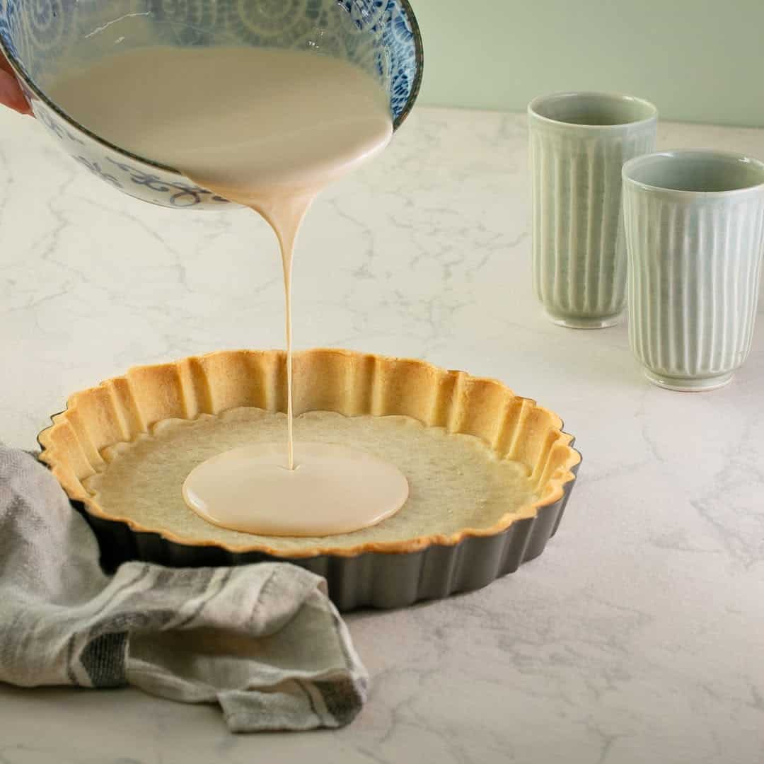 Batter being poured into a tart crust in a pan, with two cups, a cloth, and other kitchen tools for holiday baking on a light countertop.