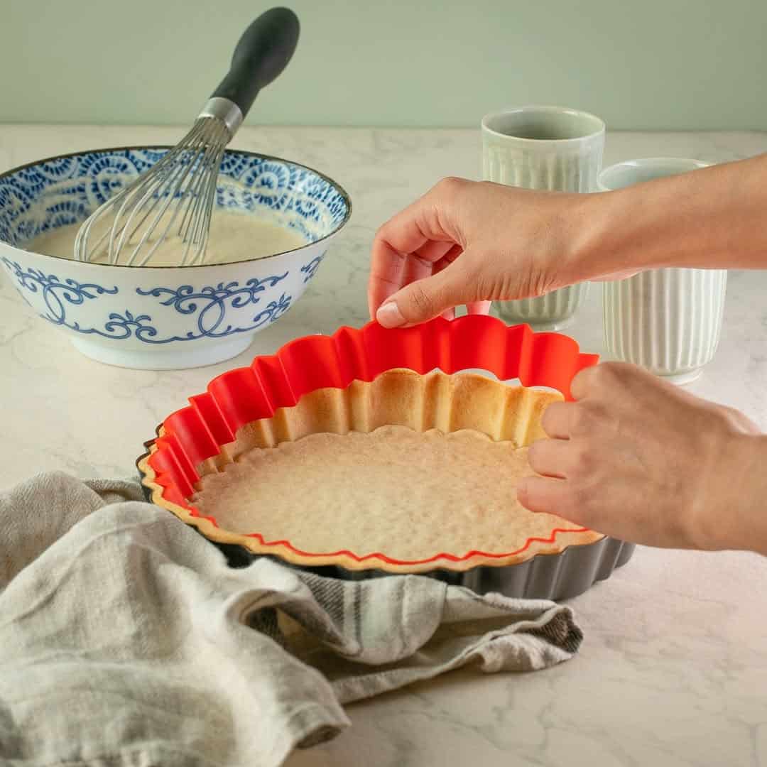 Hands removing a baked pie crust from a red silicone tart pan on a kitchen counter with baking items and essential kitchen tools for holiday baking nearby.