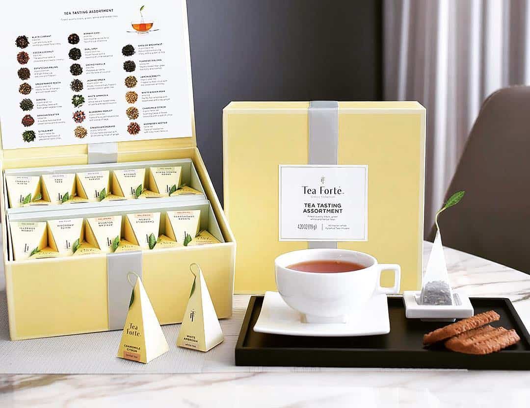 A yellow tea assortment box, a cup of tea, cookies, and a tea bag on a tray make for cozy holiday gifts set beautifully on a table.