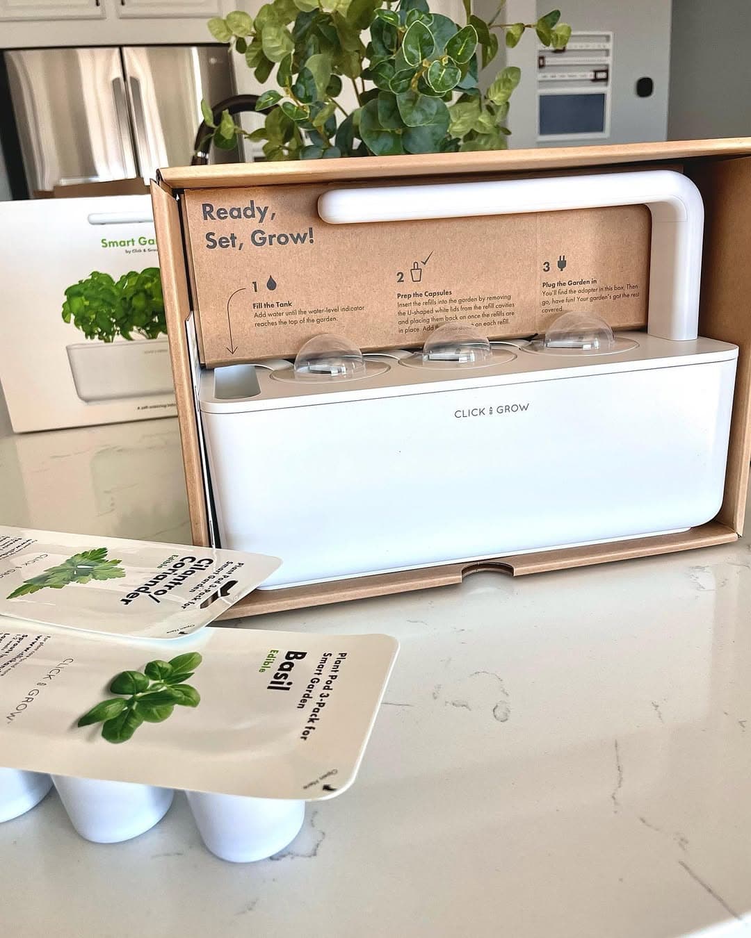 A white smart indoor garden kit with basil plant pods sits on a kitchen counter next to its packaging-an ideal gift for holiday hosts and entertainers.