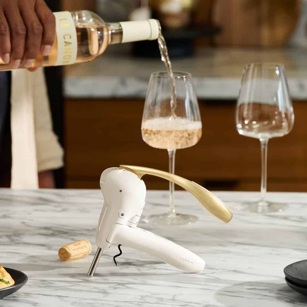 A person pours rosé wine into a glass on a marble counter with a corkscrew, two wine glasses, and some handy kitchen gadgets for holiday cooking.