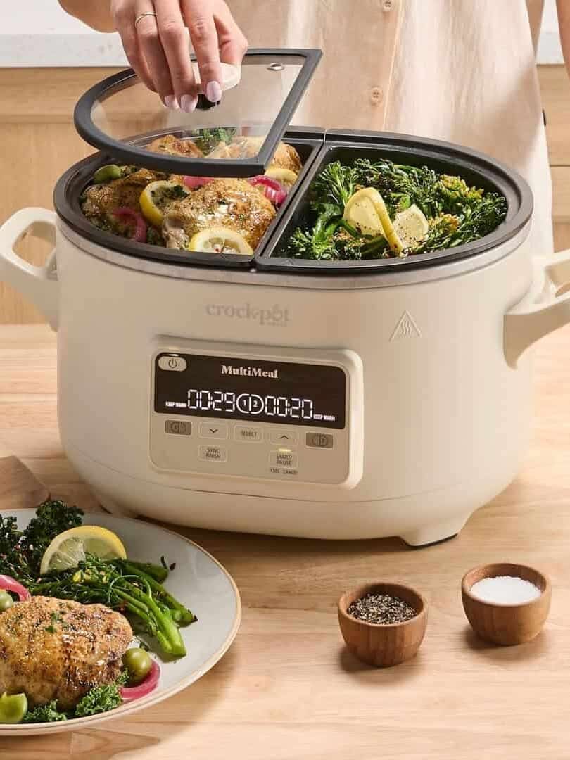 A person lifts the lid of a Crock-Pot with three sections, each holding cooked chicken, broccoli, and lemon-one of the must-have kitchen gadgets for holiday cooking.