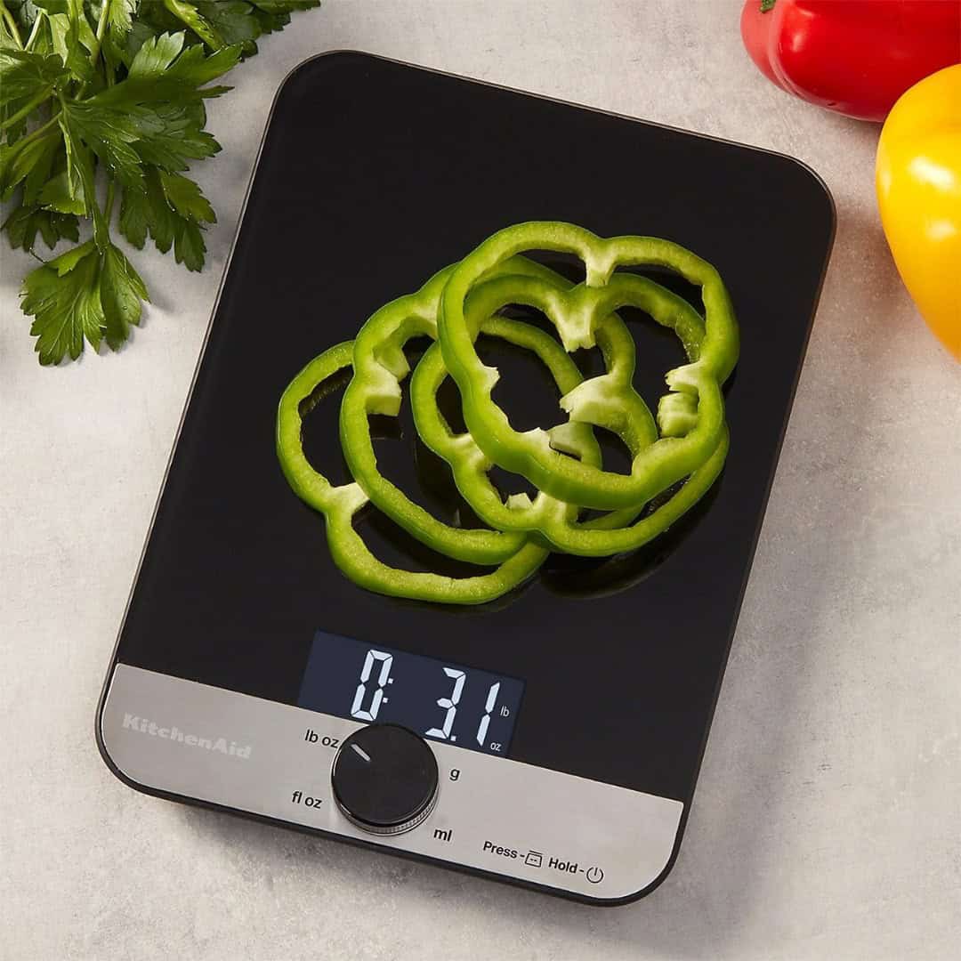 Green bell pepper slices rest on a digital kitchen scale&mdash;one of the essential kitchen tools for holiday baking&mdash;surrounded by fresh parsley, vibrant red, and yellow peppers.
