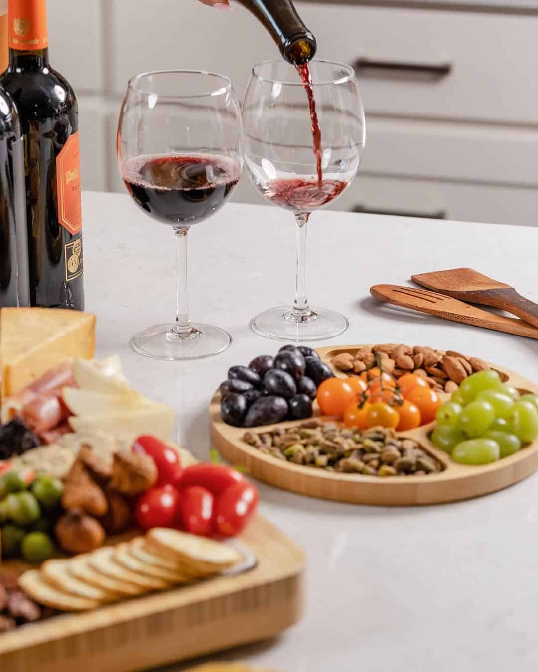 Two glasses of red wine, a bottle, fruit, nuts, cheese, and crackers arranged on a kitchen counter—perfect gifts for holiday hosts and entertainers.