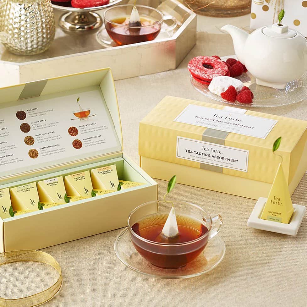 A tea tasting assortment box, teacup with pyramid tea bag, and teapot on a beige tablecloth—perfect gifts for holiday hosts and entertainers.