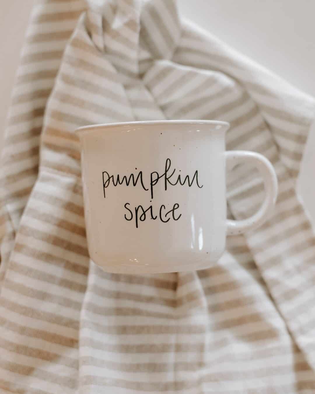 White mug with "pumpkin spice" written on it, placed on a beige and white striped cloth background—perfect for anyone who loves unique pumpkin spice gifts.