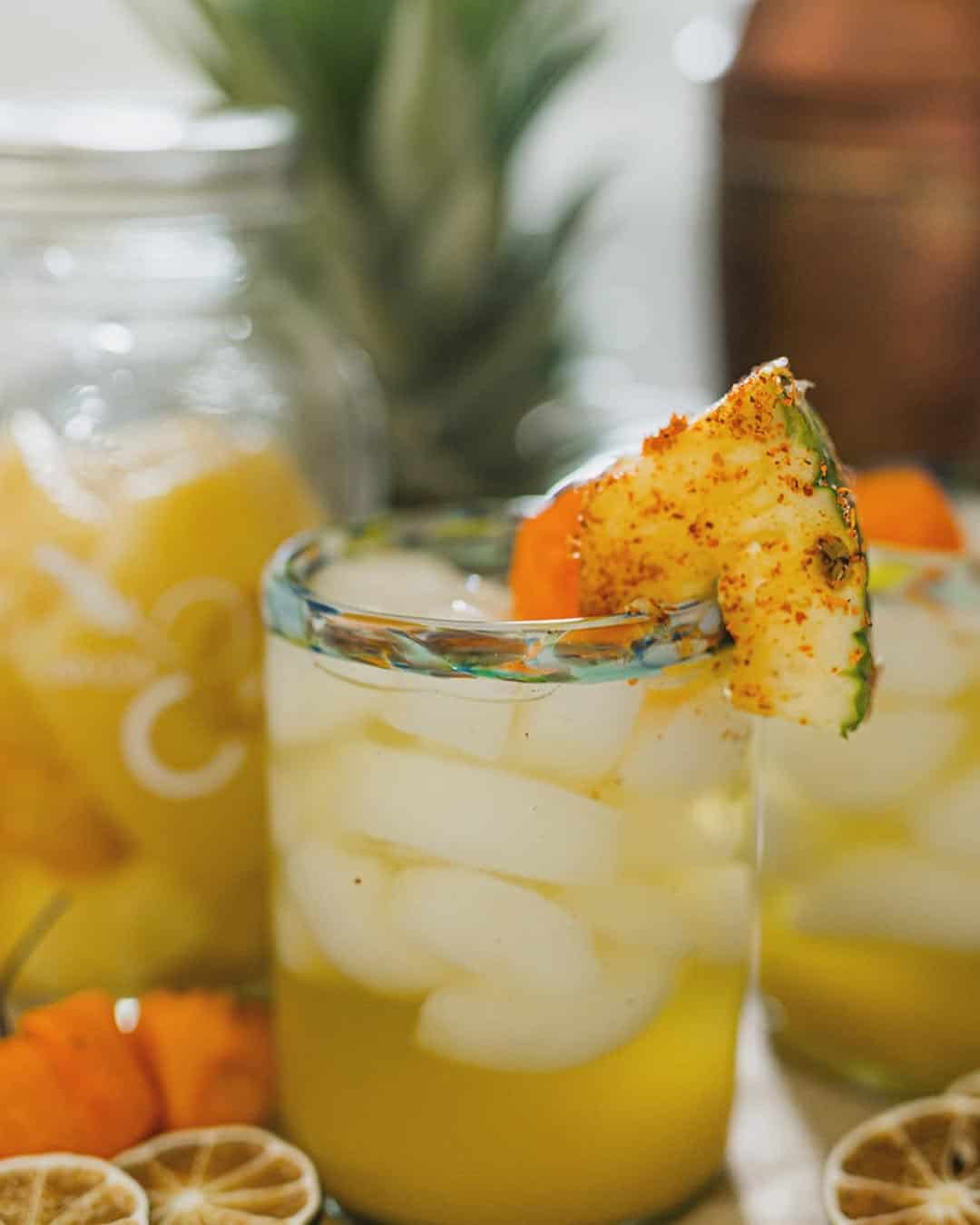 A glass of iced yellow drink garnished with pineapple and chili powder sits front and center, perfect as a refreshing idea for Gifts for Holiday Hosts and Entertainers, with colorful fruit jars in the background.