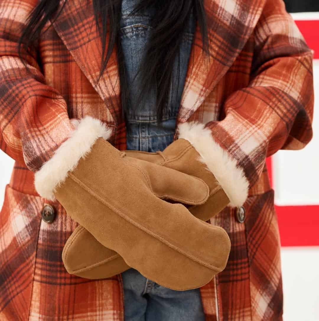 Person in a plaid coat and denim shirt crossing their arms, wearing tan mittens with white fur trim—perfect inspiration for cozy holiday gifts.