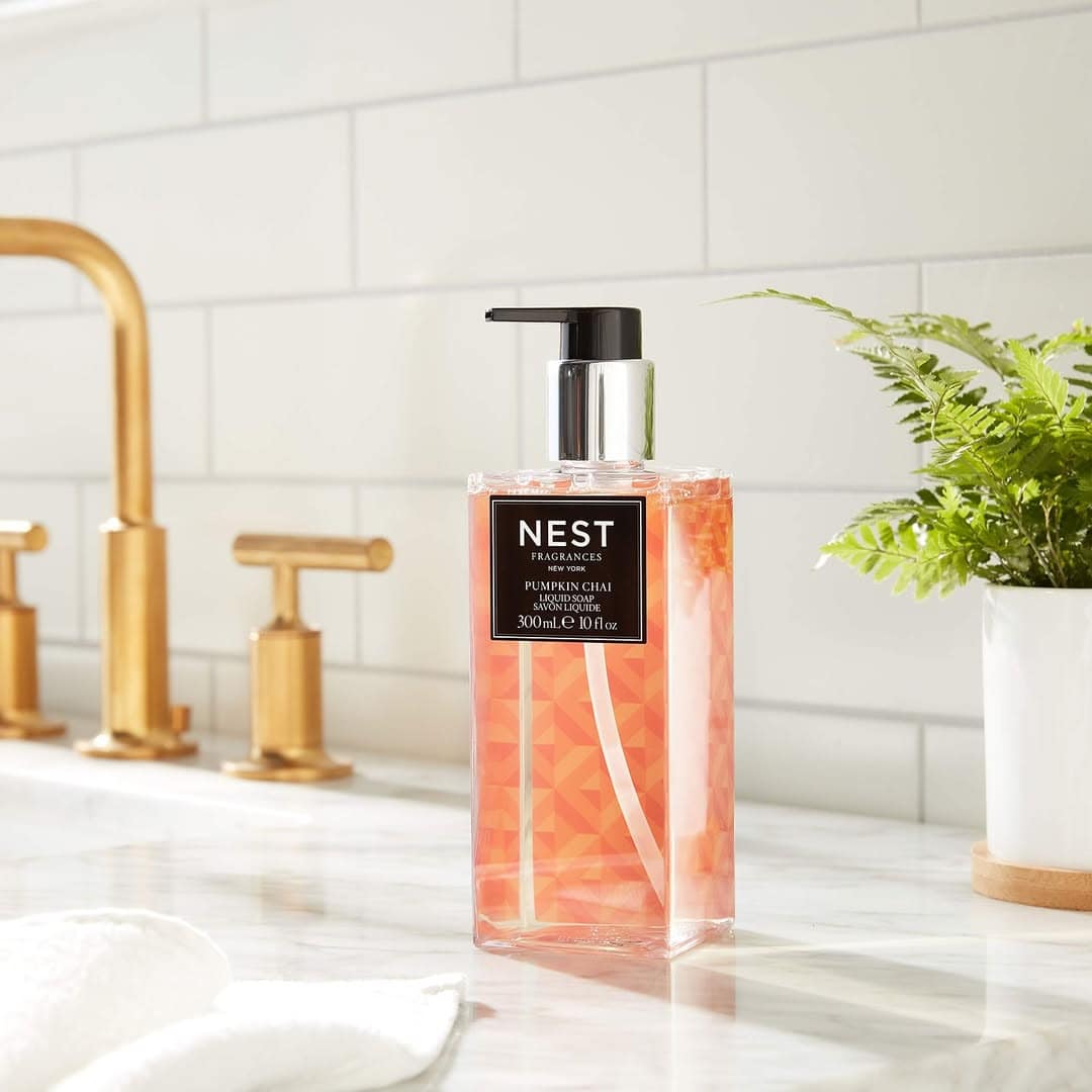 A bottle of NEST Pumpkin Chai hand soap, the perfect addition to your pumpkin spice gifts, sits on a marble counter by a sink with gold fixtures and a potted plant.