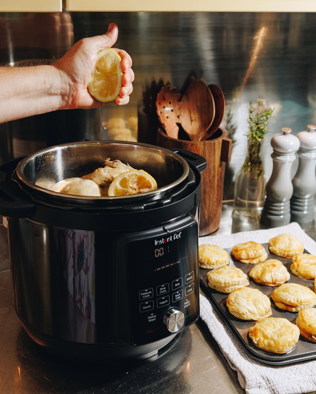 A hand squeezes a lemon over an Instant Pot—one of the must-have kitchen gadgets for holiday cooking—while baked pastries cool nearby in a cozy kitchen.