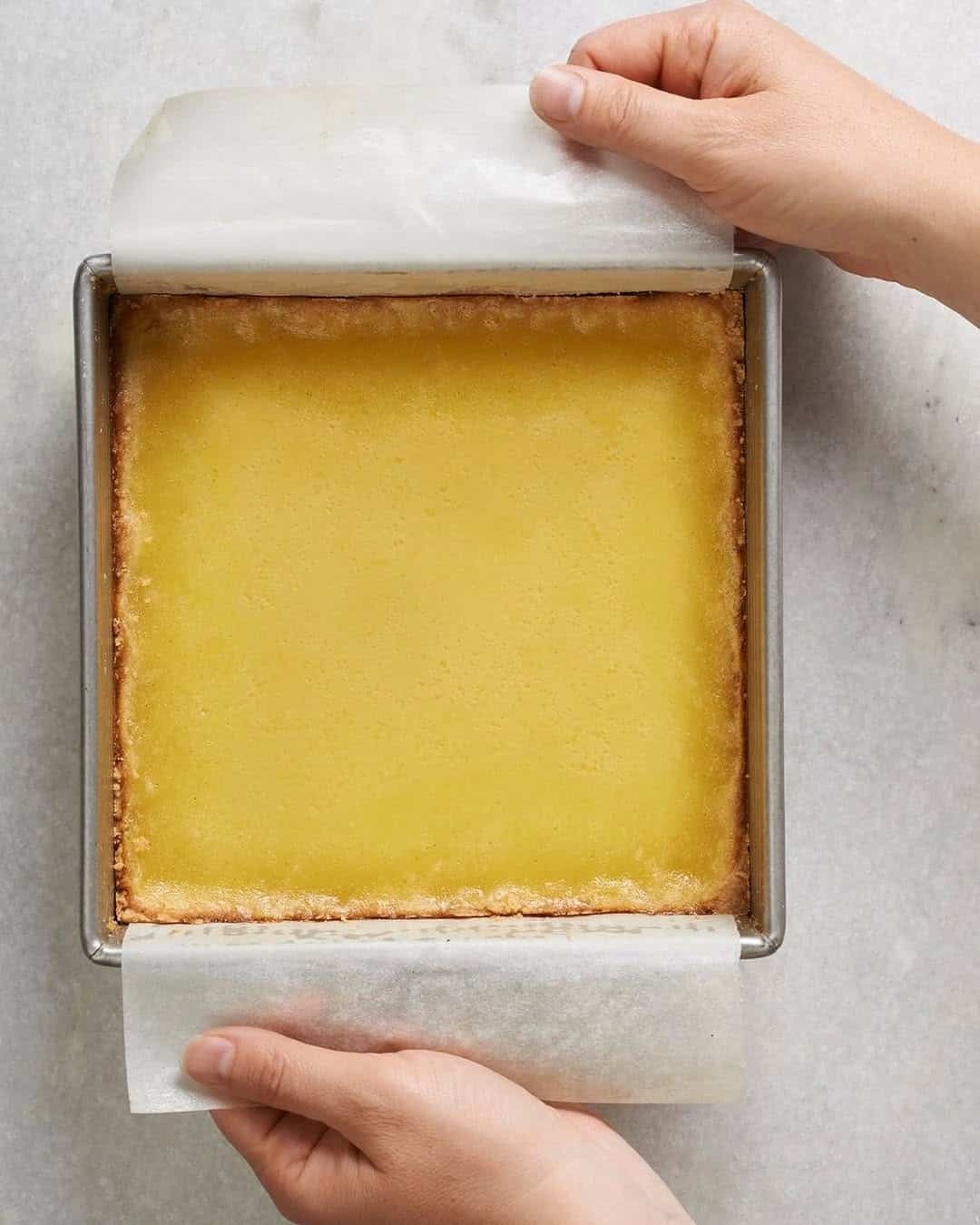 Hands lifting lemon bars from a square baking pan using parchment paper on a light countertop&mdash;showcasing essential kitchen tools for holiday baking.