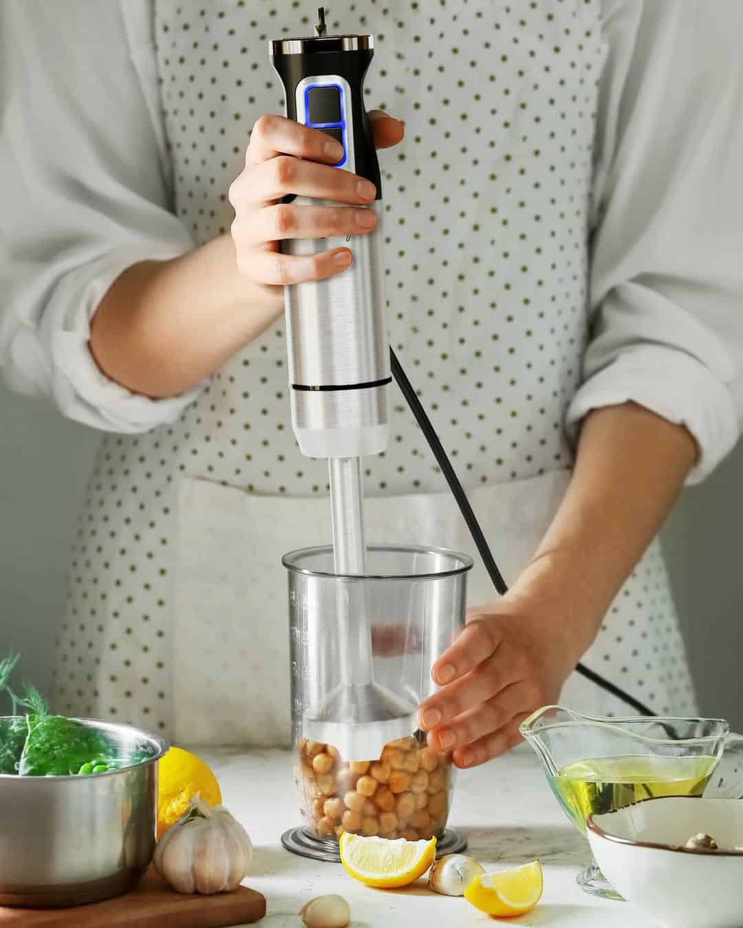 A person uses an immersion blender to blend chickpeas in a container, surrounded by fresh ingredients—a perfect example of smart kitchen gadgets for holiday cooking.