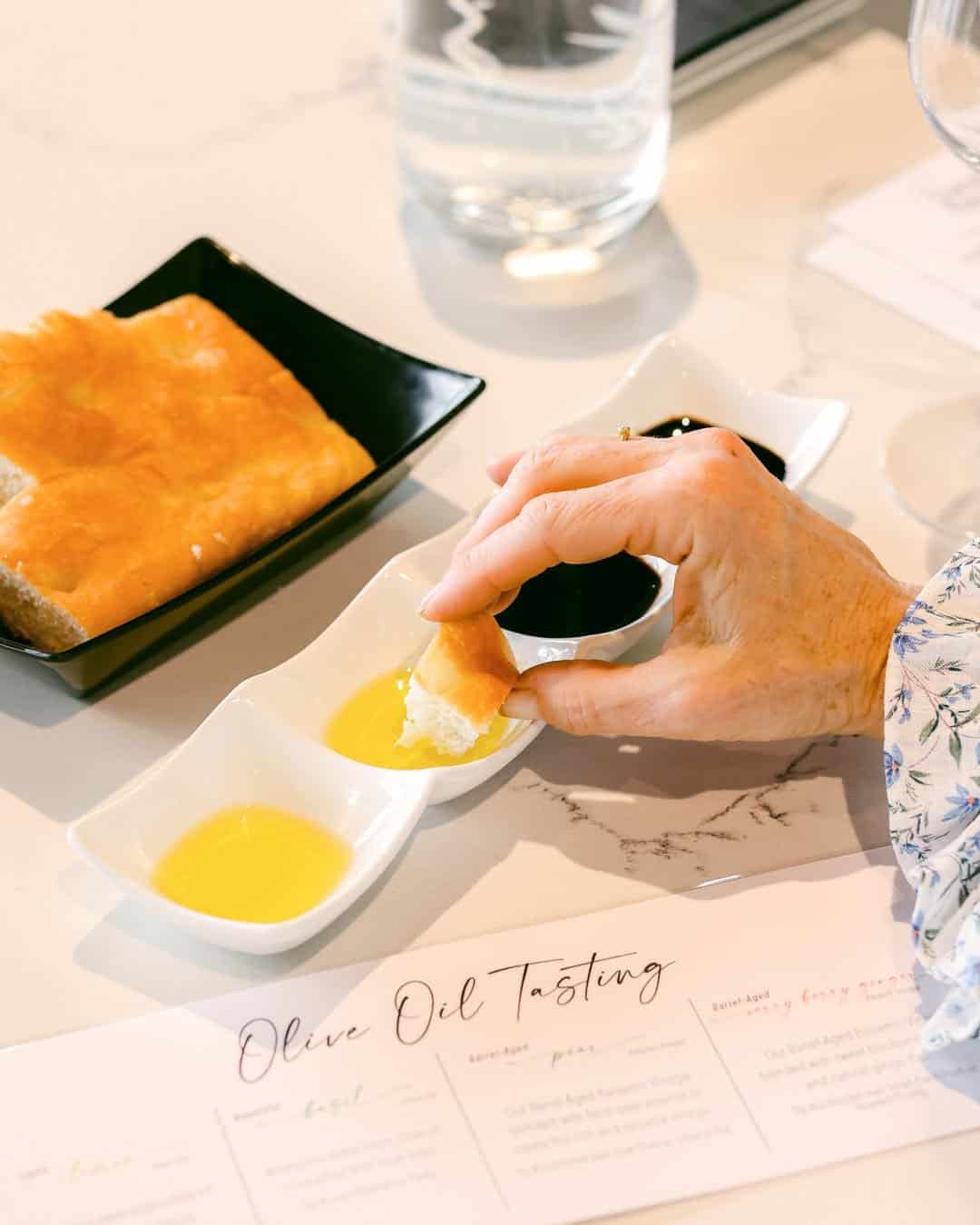 Hand dipping bread into olive oil, surrounded by tasting dishes, bread, and an "Olive Oil Tasting" menu-perfect gifts for holiday hosts and entertainers.