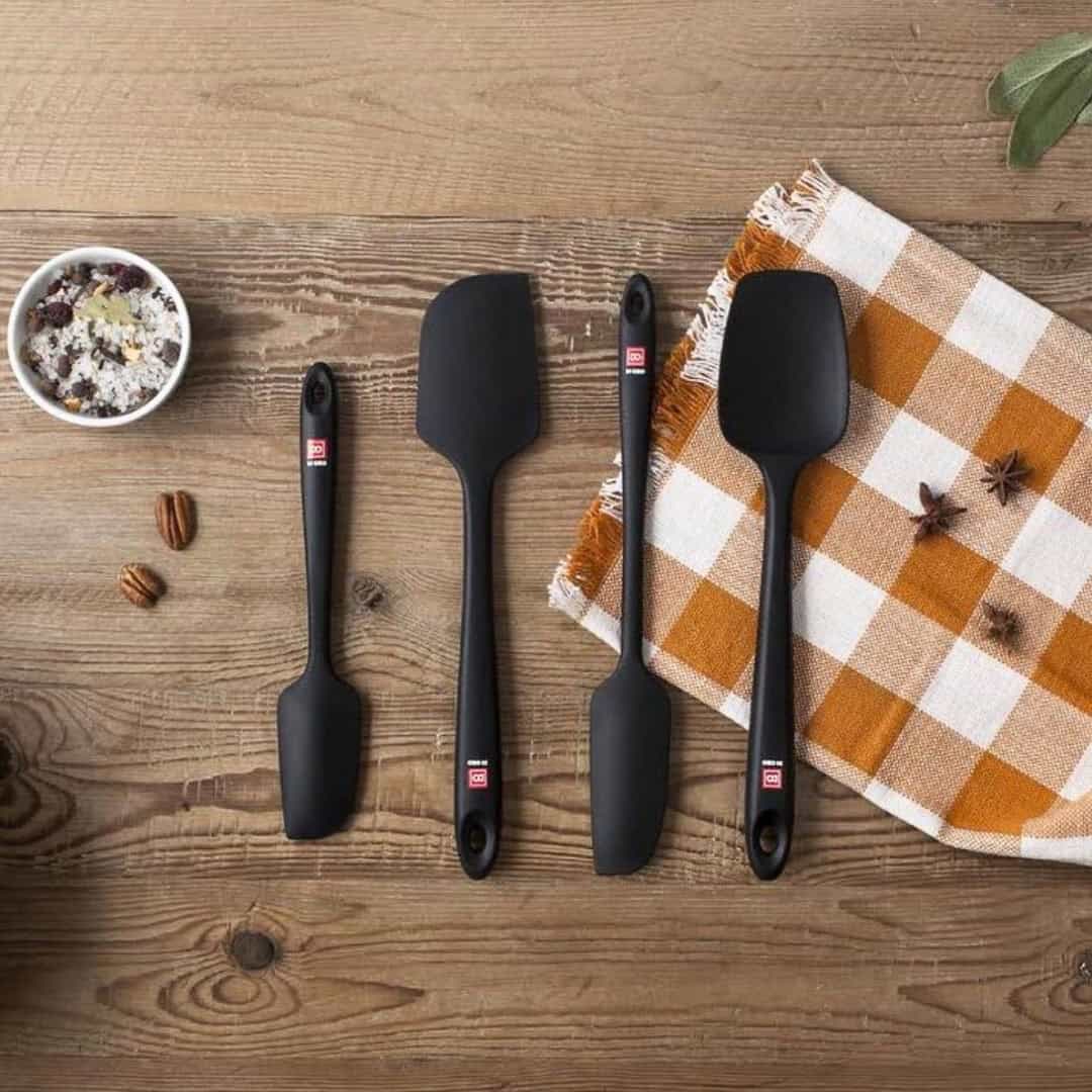 Four black spatulas, perfect kitchen gadgets for holiday cooking, rest on a wooden table beside a checkered cloth and a bowl of spices.