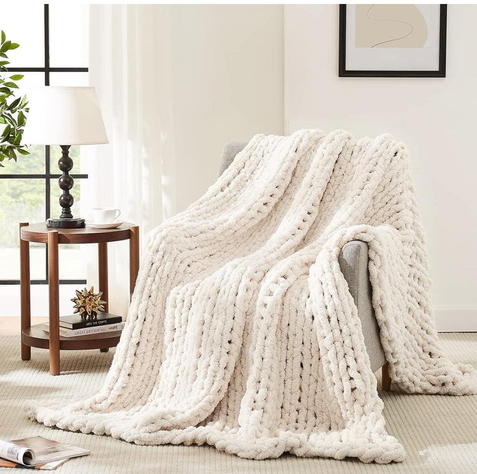 Cream-colored chunky knit blanket draped over a gray armchair in a cozy, well-lit living room—perfect for those seeking cozy holiday gifts.