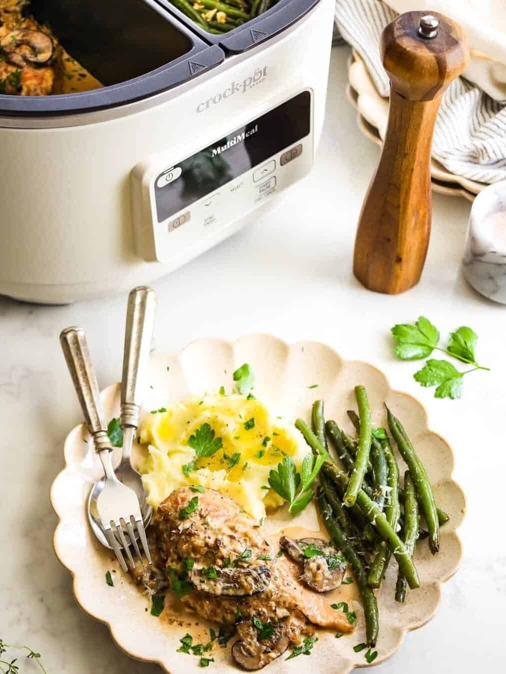 Chicken with mashed potatoes and green beans on a plate, next to a Crockpot, pepper grinder, parsley, and essential kitchen gadgets for holiday cooking.