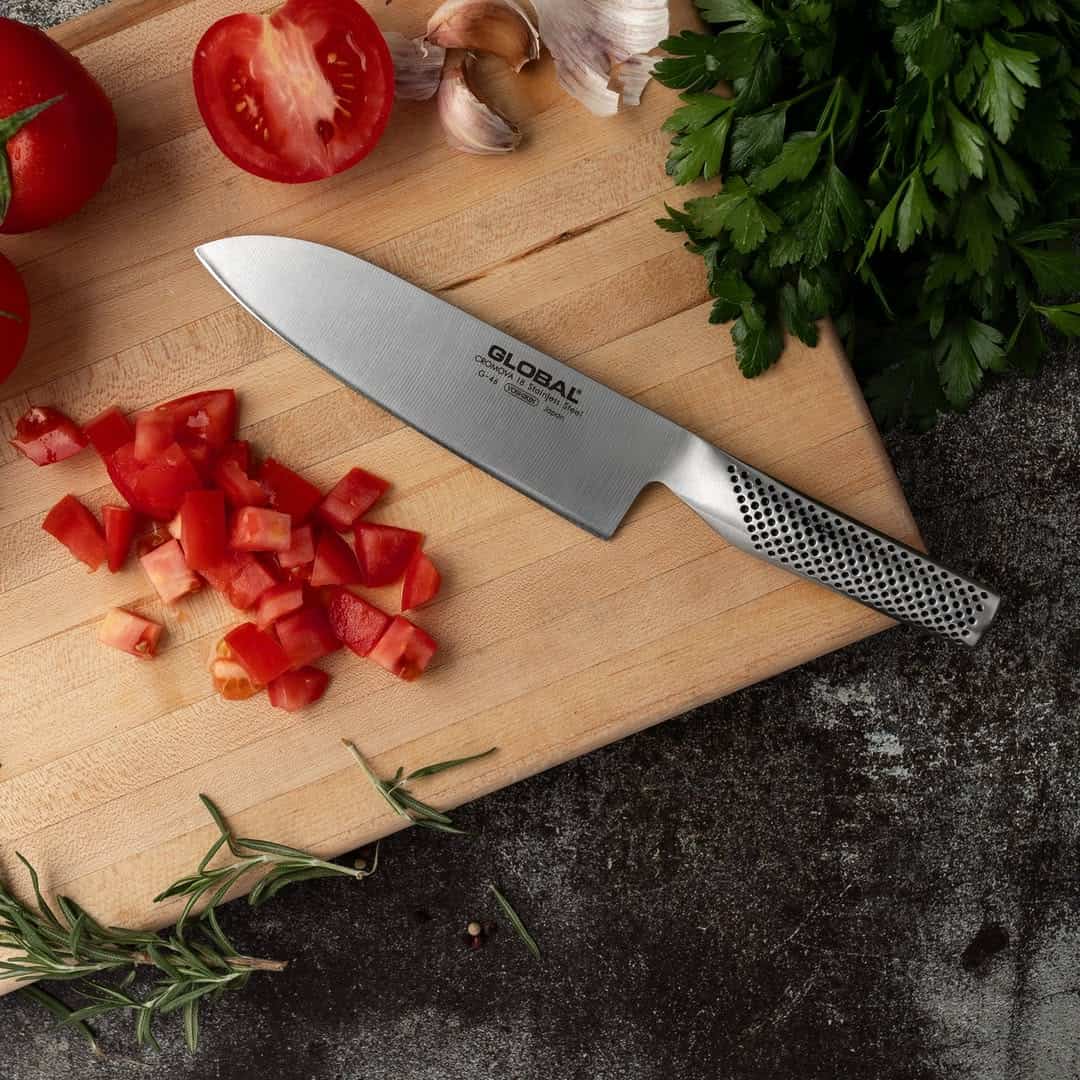 A chef’s knife on a cutting board with chopped tomatoes, whole tomatoes, parsley, garlic, and rosemary—essential kitchen gadgets for holiday cooking.
