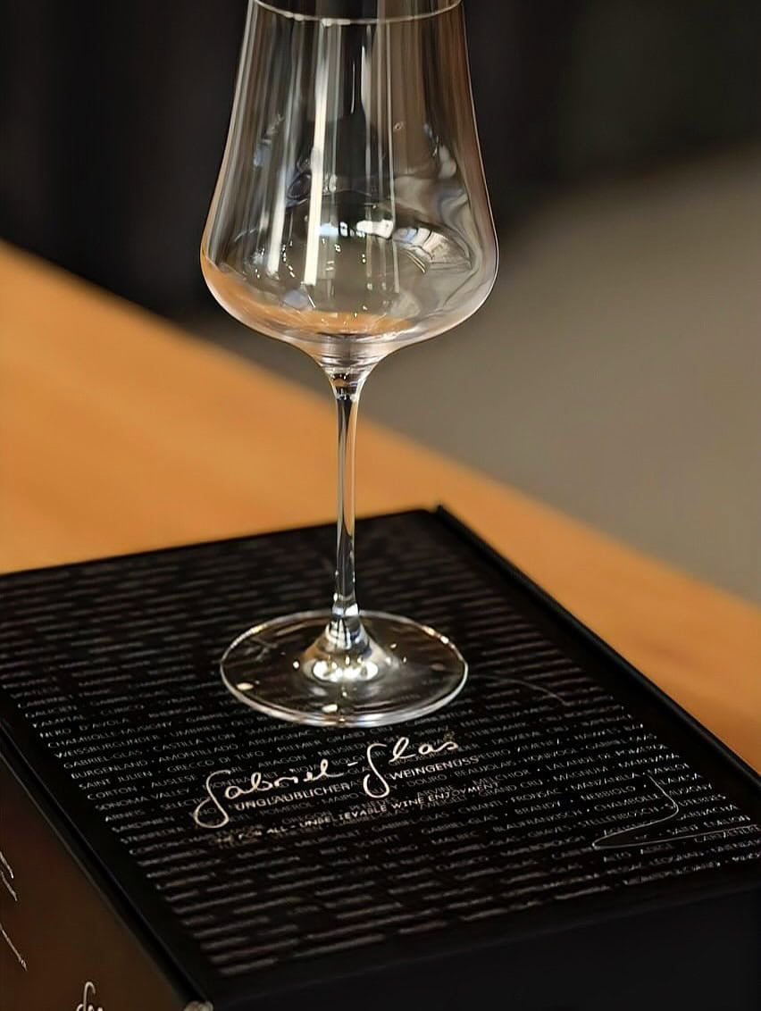A clear wine glass sits on top of a black box with white script writing on its lid-an elegant choice among gifts for holiday hosts and entertainers.