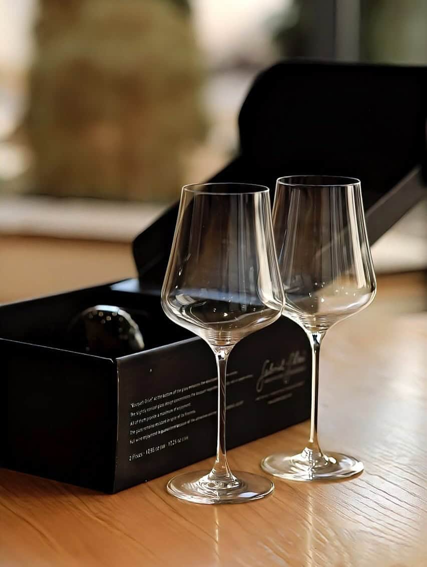 Two empty wine glasses stand next to an open black box on a wooden table, making elegant gifts for holiday hosts and entertainers.
