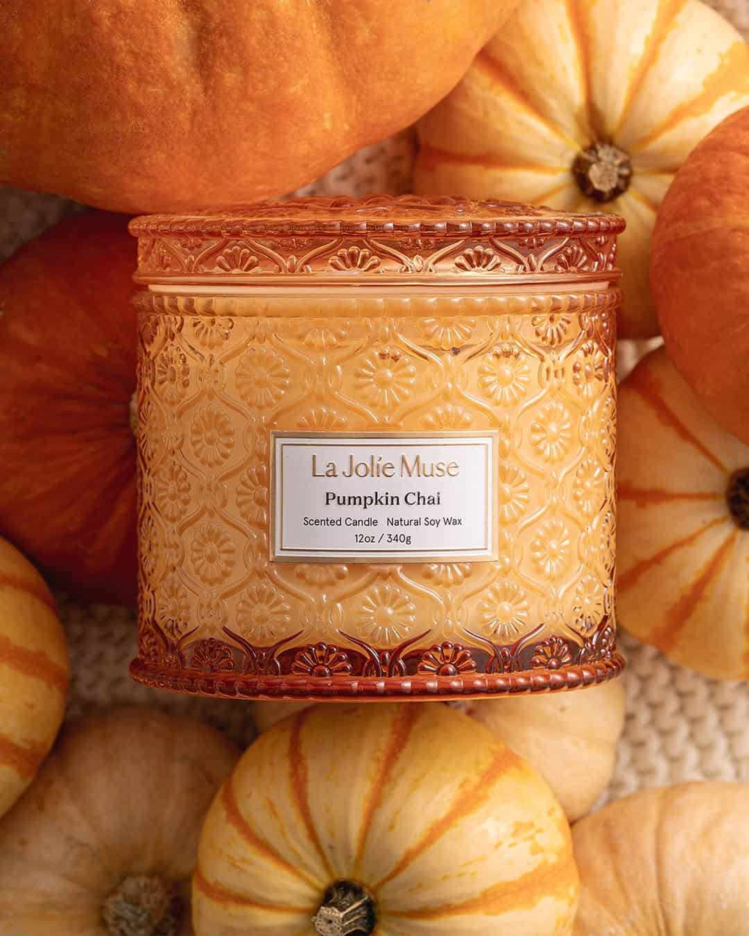 Ornate orange candle labeled "Pumpkin Chai" surrounded by small orange and white pumpkins.