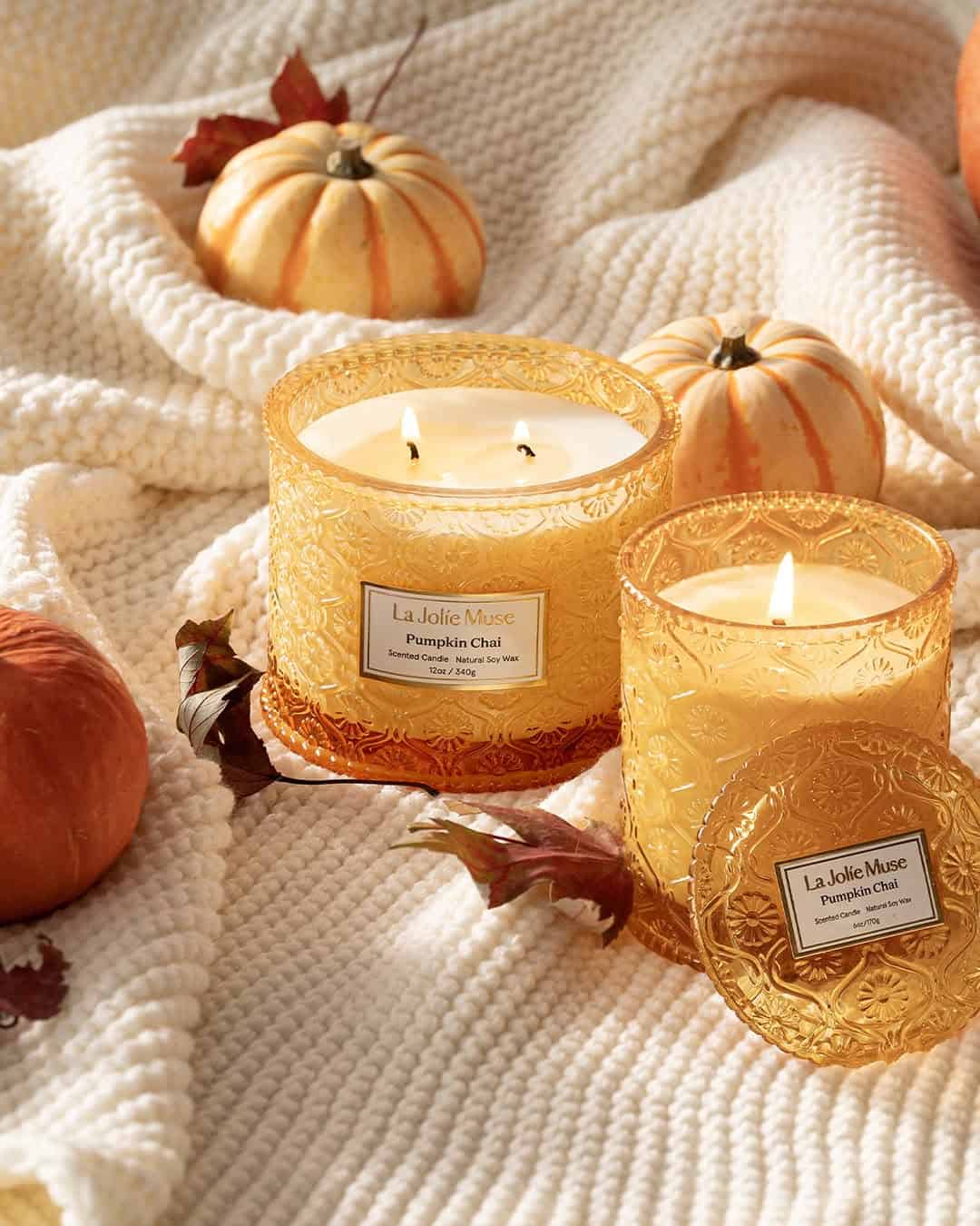 Two lit pumpkin chai candles on a cream knit blanket, surrounded by mini pumpkins and autumn leaves.