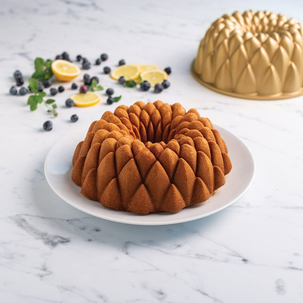 A decorative bundt cake on a white plate, topped with blueberries and lemon slices, sits beside a gold bundt pan-showcasing essential kitchen tools for holiday baking.
