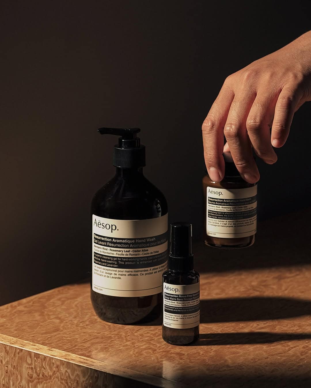 A hand reaches for one of three Aesop skincare bottles on a wooden surface, lit by warm light—an ideal gesture for those seeking thoughtful gifts for holiday hosts and entertainers.
