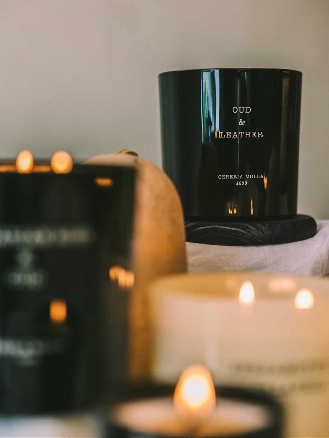 Black candle labeled "Oud & Leather" surrounded by other lit candles with a soft, warm glow-perfect for adding ambiance to your space or as cozy holiday gifts.