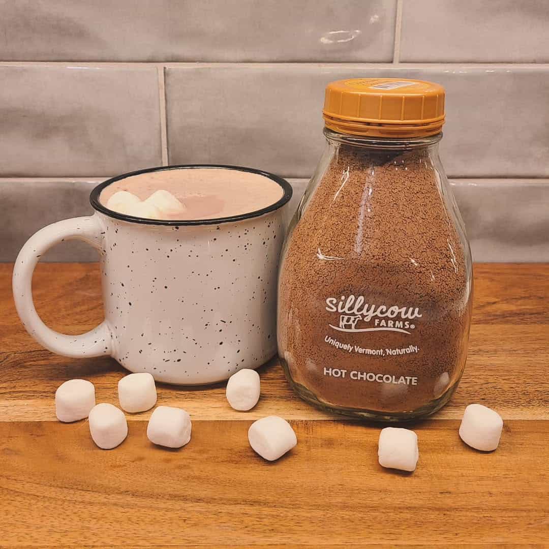 A mug of hot chocolate with marshmallows and a jar of Sillycow Farms hot chocolate sit on a wooden surface—perfect cozy holiday gifts.