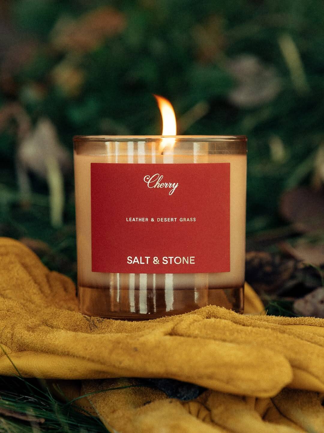 A lit candle labeled “Cherry, Salt & Stone” sits on yellow gloves with grass and leaves in the background—an elegant choice among gifts for holiday hosts and entertainers.