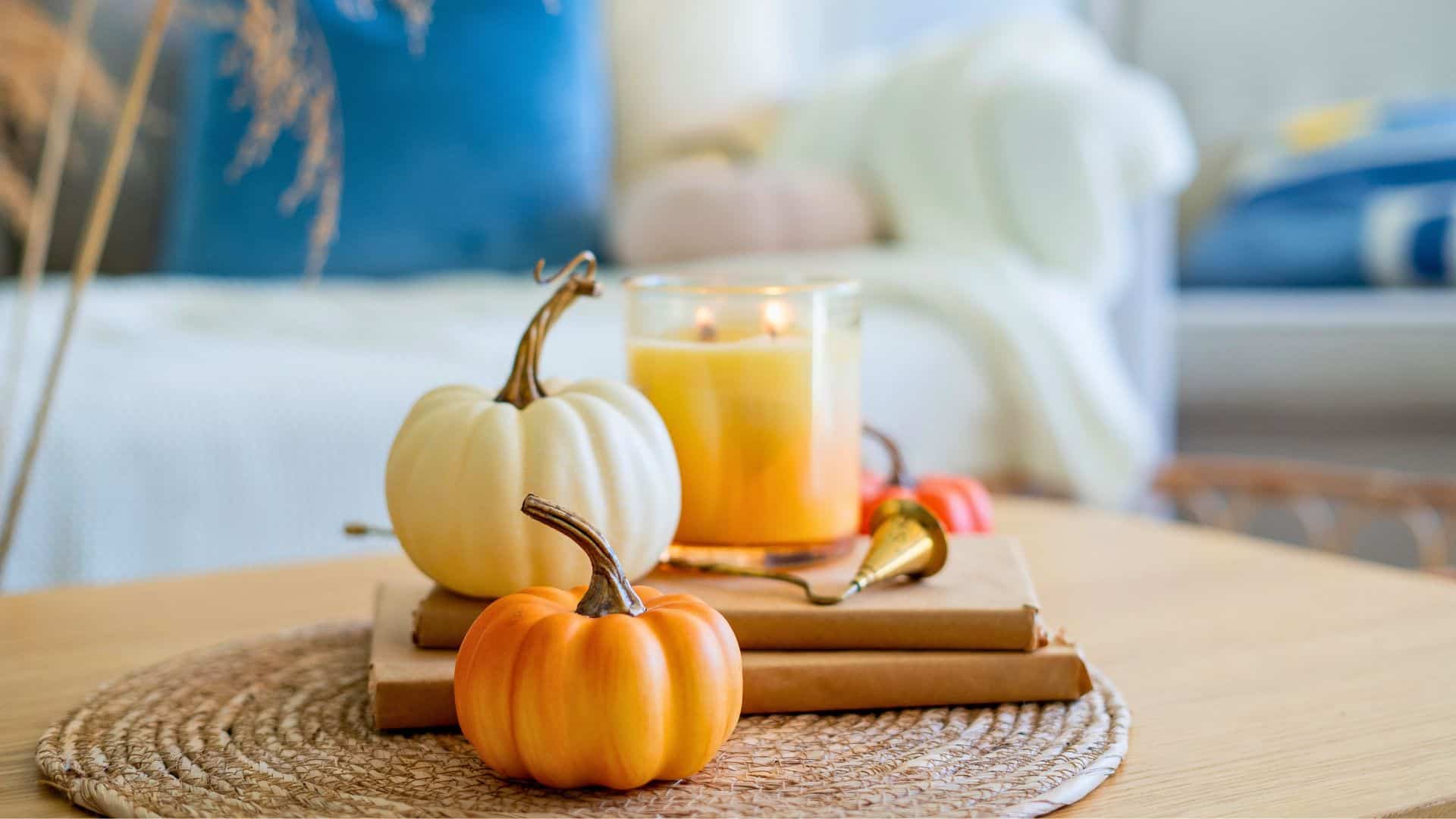 White and orange pumpkins, a lit candle, books, and pumpkin spice gifts on a woven placemat create a cozy autumn scene.