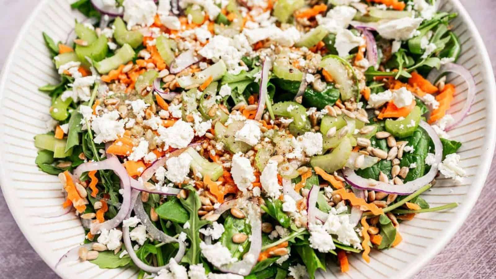 A salad with leafy greens, feta cheese, shredded carrots, onions, sunflower seeds, and dressing on a plate.