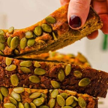 A hand picking up a slice of pumpkin bread topped with pumpkin seeds from a stacked loaf.