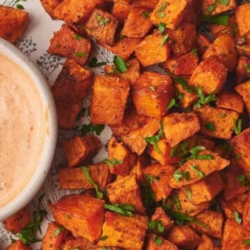 Roasted sweet potato cubes garnished with herbs on a plate, next to a bowl of creamy dipping sauce.