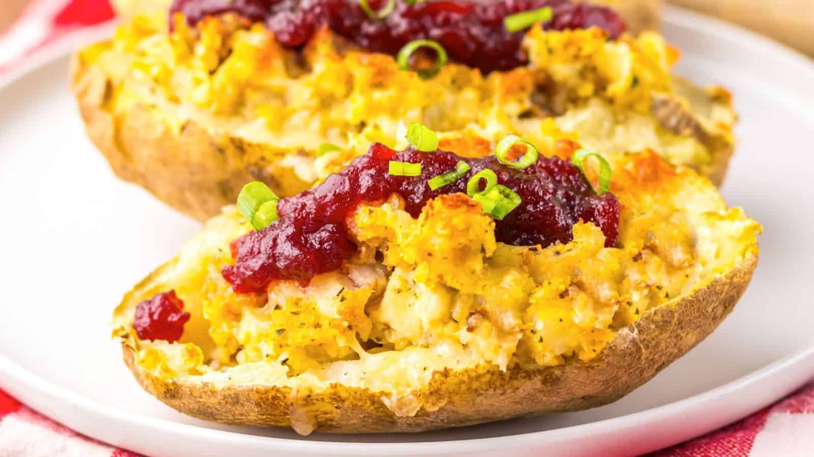 A close-up of Thanksgiving-inspired potato skins, featuring two baked potatoes loaded with mashed potato filling, leftover turkey and stuffing, twice baked potatoes,and topped with cranberry sauce, and chopped green onions, served on a white plate.