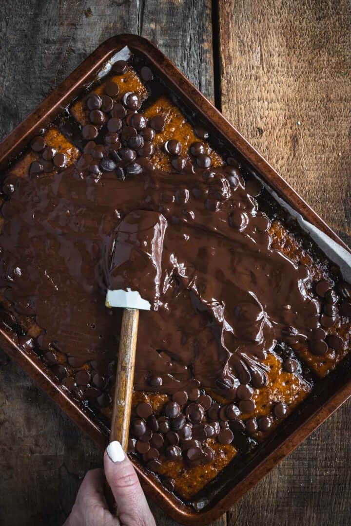 A hand spreads melted chocolate over a rectangular cake with a spatula on a rustic wooden table, creating a look reminiscent of Christmas Toffee Bark.