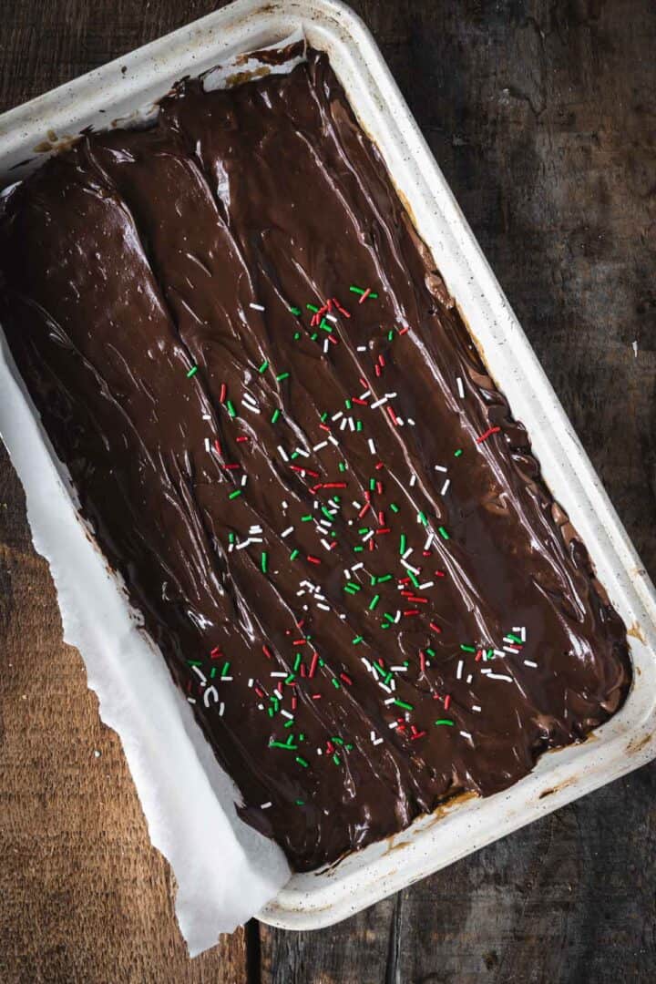 A tray of Christmas Toffee Bark fudge topped with red, green, and white sprinkles sits on a rustic wooden table.