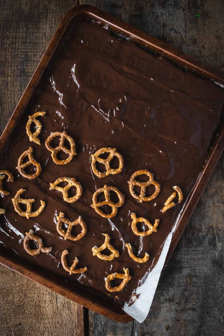 A baking tray with melted chocolate and pretzels arranged on top, set on a rustic wooden surface&mdash;perfect for making festive Christmas Toffee Bark.