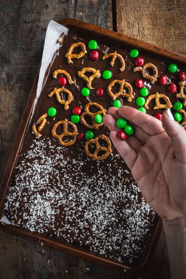 A hand sprinkles red and green M&Ms onto Christmas Toffee Bark with pretzels and shredded coconut on a baking tray.
