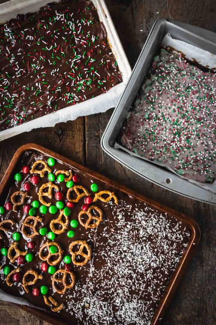 Three pans of Christmas Toffee Bark topped with sprinkles, pretzels, candies, and coconut rest on a wooden surface.