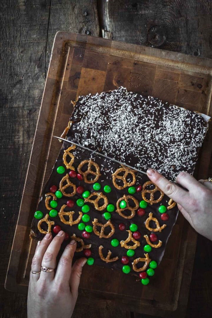Hands cutting Christmas Toffee Bark topped with pretzels, M&Ms, and coconut on a wooden board.
