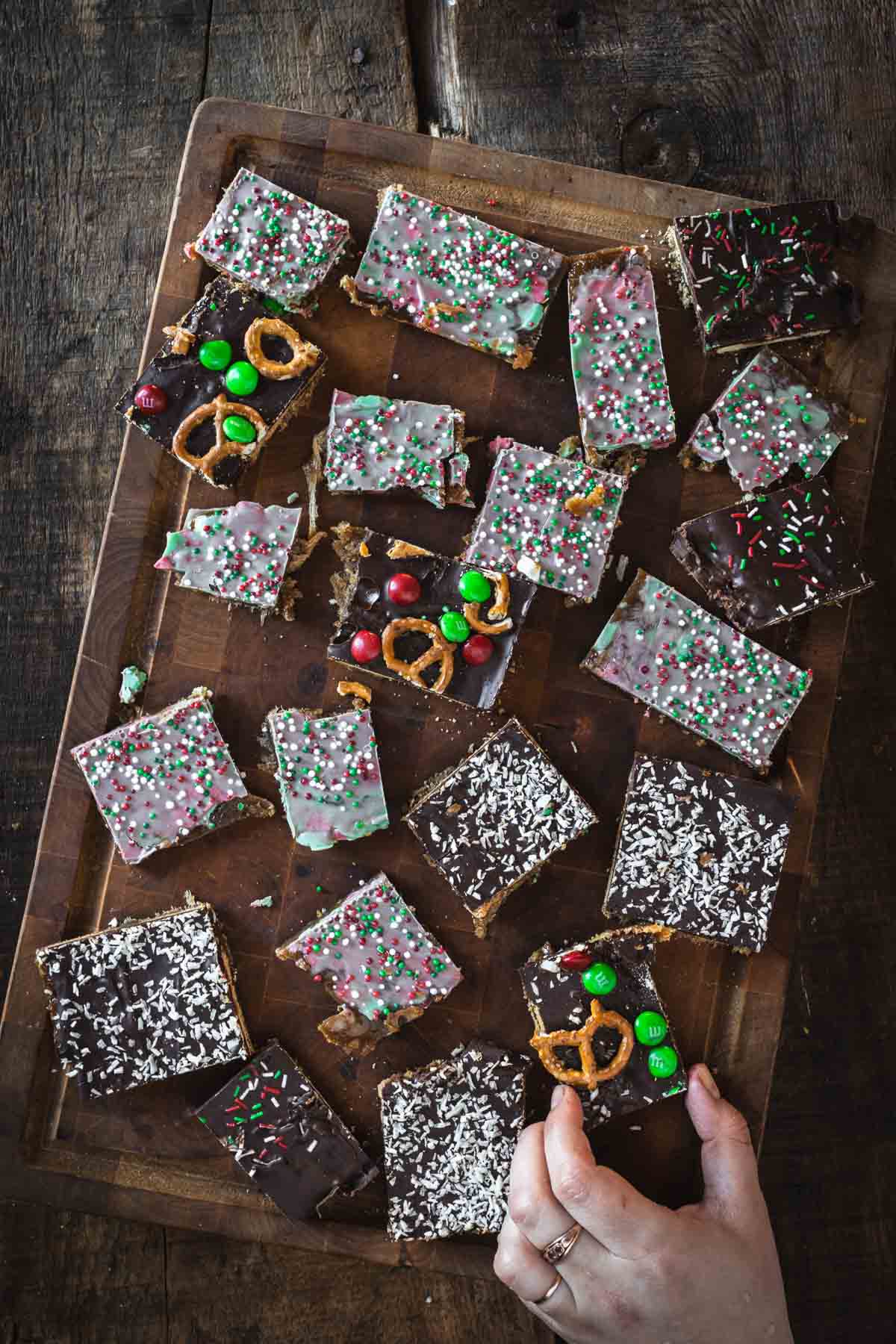 A hand reaches for a Christmas Toffee Bark, a chocolate-topped dessert bar, on a wooden board with various decorated bars.