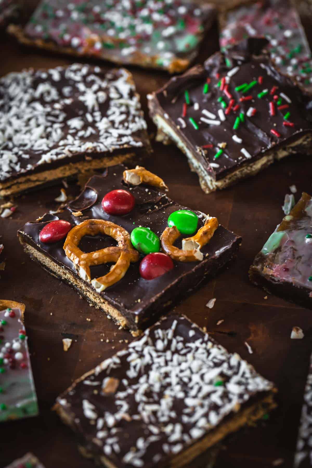 Christmas Toffee Bark bars topped with pretzels, M&Ms, coconut flakes, and sprinkles are artfully arranged on a dark surface.