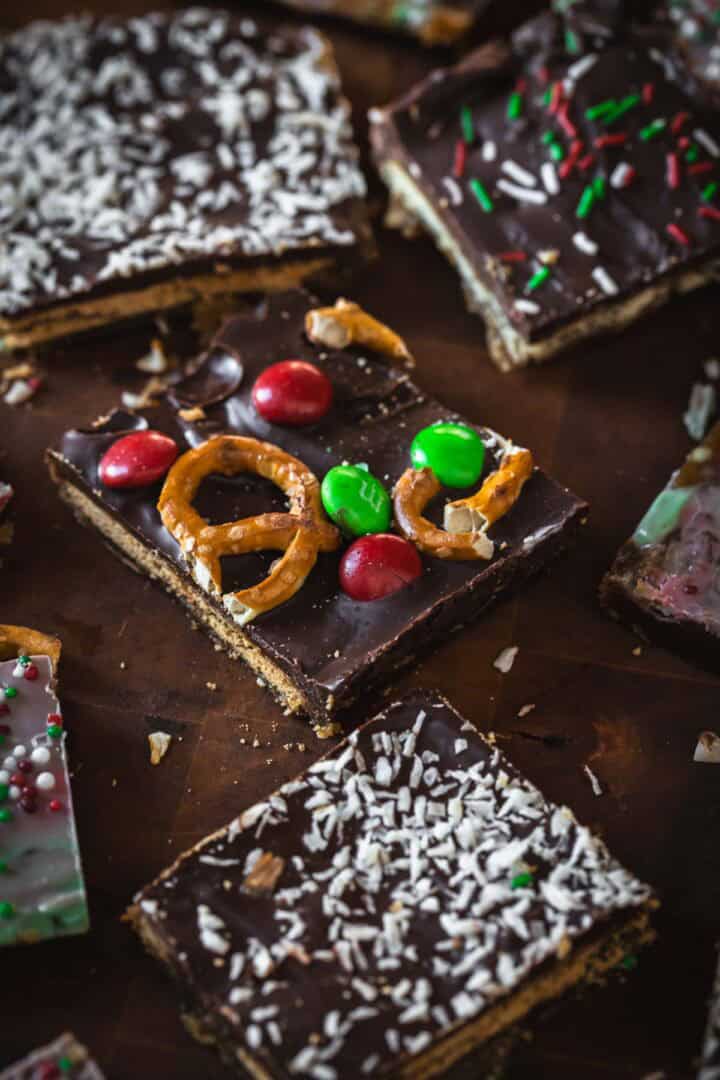 Christmas Toffee Bark squares topped with pretzels, red and green candies, coconut flakes, and sprinkles are displayed on a dark surface.