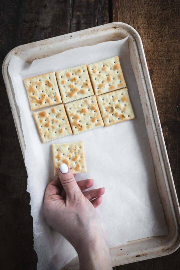 A hand holding a square cracker above a tray of baked crackers on parchment paper, perfect for making Christmas Toffee Bark.
