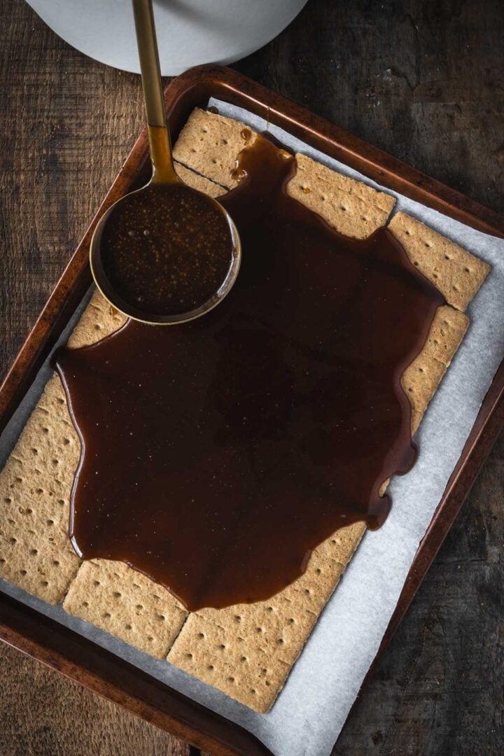 A ladle pours dark syrup over graham crackers on a parchment-lined baking tray, the first step in creating festive Christmas Toffee Bark.