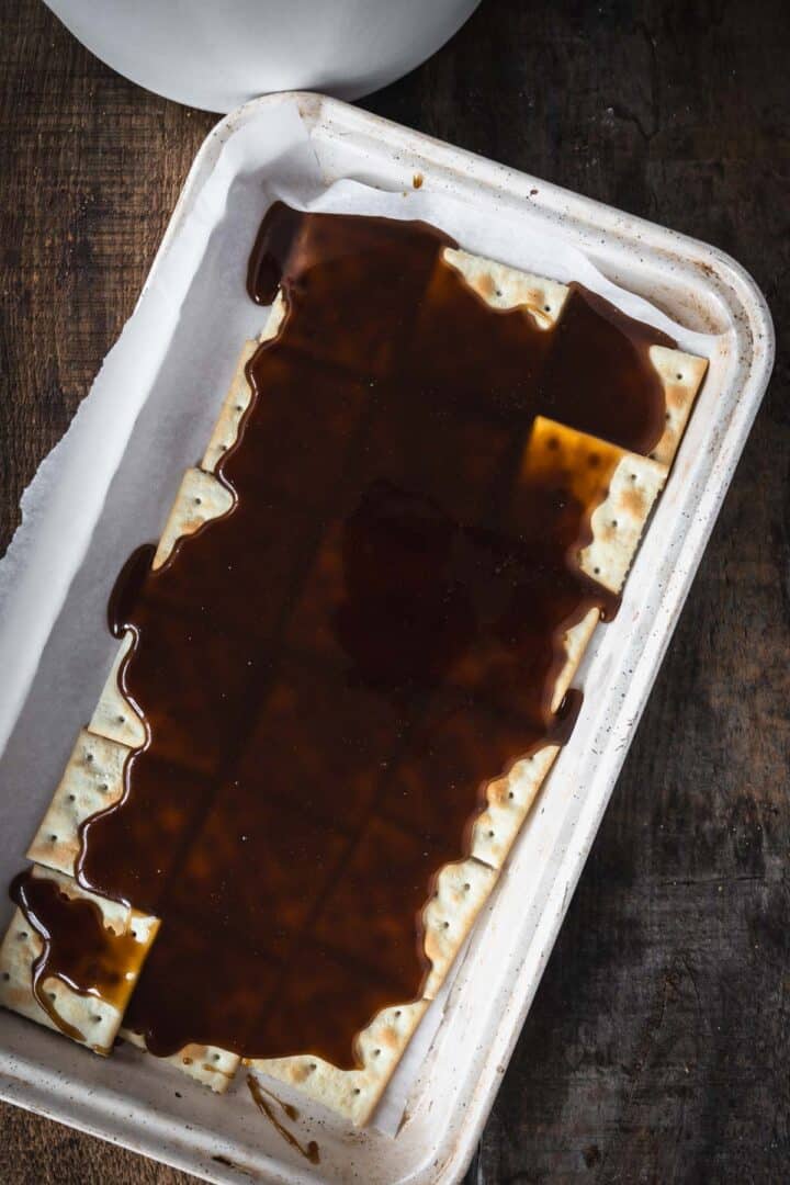 Matzo crackers on a tray are covered with a glossy layer of dark syrup, creating a treat reminiscent of Christmas Toffee Bark, set on a rustic wooden surface.