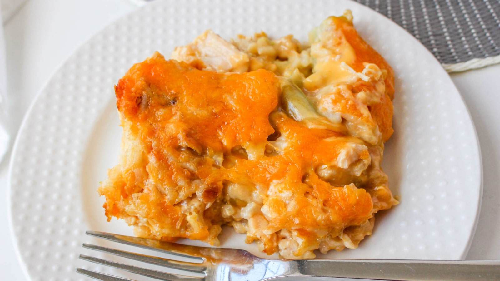 A serving of cheesy baked casserole with chicken and vegetables on a white plate. A fork is placed beside the dish.
