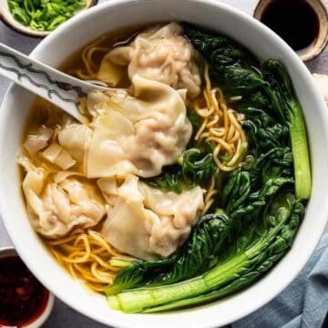 Bowl of wonton noodle soup with greens, surrounded by sauces, garlic, ginger, and chopsticks.
