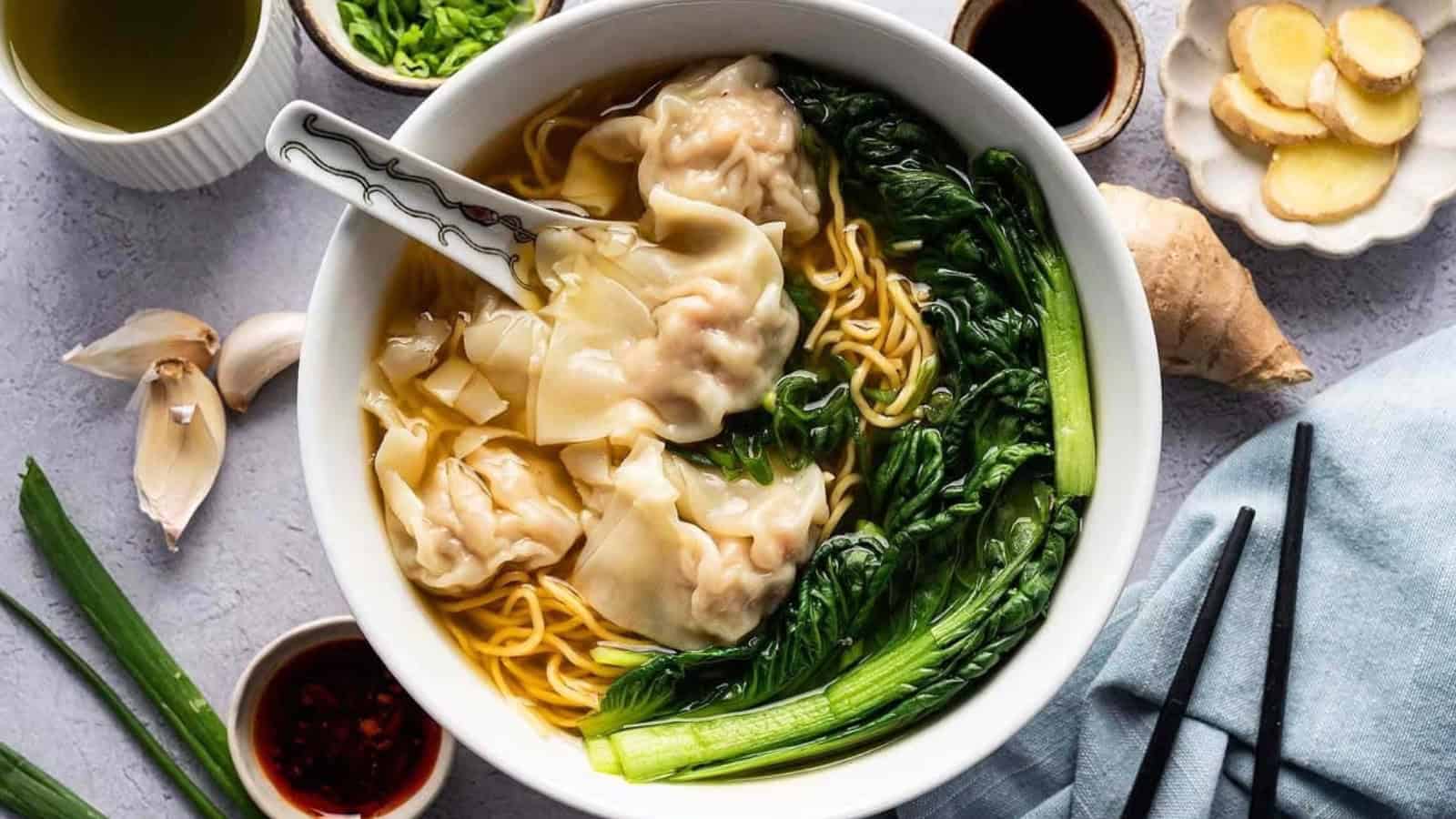 Bowl of wonton noodle soup with greens, surrounded by sauces, garlic, ginger, and chopsticks.